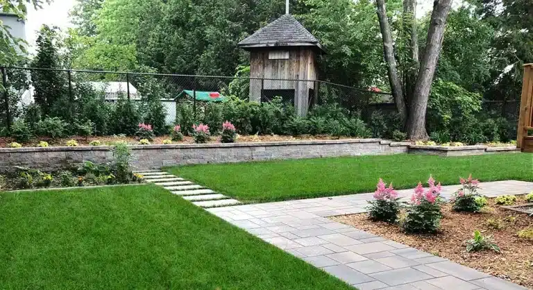 Aménagement paysager résidentiel avec mur de soutènement en blocs de béton, pelouse verdoyante, allée pavée et plates-bandes fleuries.